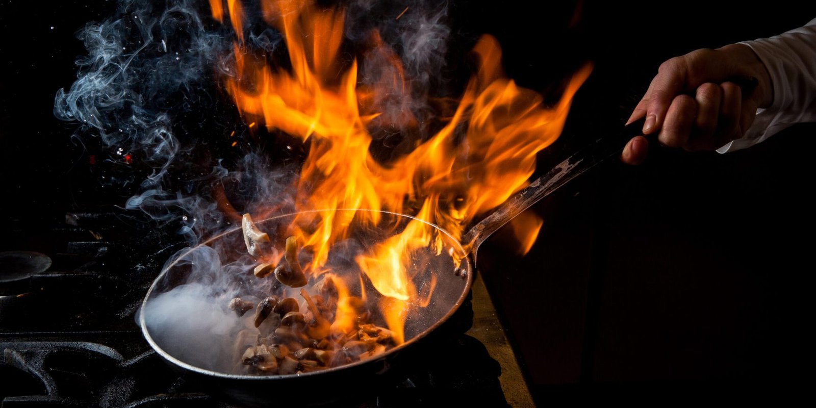side view mushroom frying with stove and fire and human hand in pan on black background